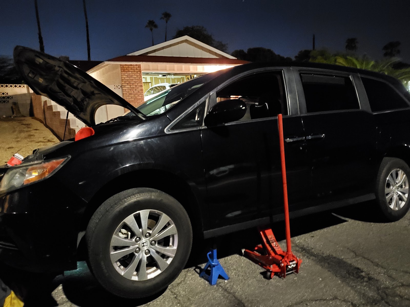 Black Honda Odyssey minivan with hood open, raised on jack stands in a driveway at dusk